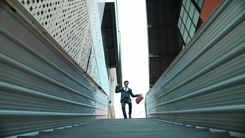 Low angle view of young smiling business man dance between building at center point. Skilled executive manager wearing suit and suitcase and moving to music. Happy investor moving to music. Exultant.