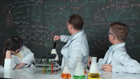 Creative teacher pointing chemistry at blackboard while talking to high school boy at table with microscope and test tube with colored liquid. Young student wearing lab coat in STEM class. Edification - Powered by Shutterstock - Get 15% off with code: PIKWIZARD15