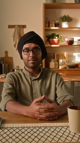 Vertical medium portrait of smiling young adult Indian man in glasses, traditional black turban and khaki kurta sitting at dining table in modern domestic kitchen and looking at camera