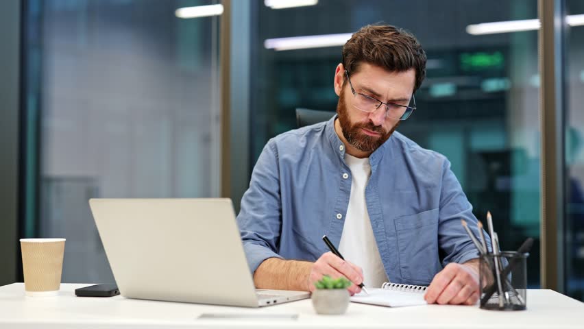 Smiling businessman writes in notebook at modern office desk. Laptop, smartphone, coffee, pens and pencils nearby. Busy professional business man company employee, young male worker software developer