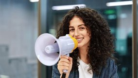Curly haired woman makes an announcement with megaphone in an office. She is smiling and looks happy to share the news. - Powered by Shutterstock - Get 15% off with code: PIKWIZARD15