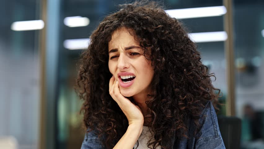Unhappy curly haired businesswoman suffering from toothache in modern office. Stressed female manager touching cheek and head, feeling painful tooth throbbing or teeth sensitivity.