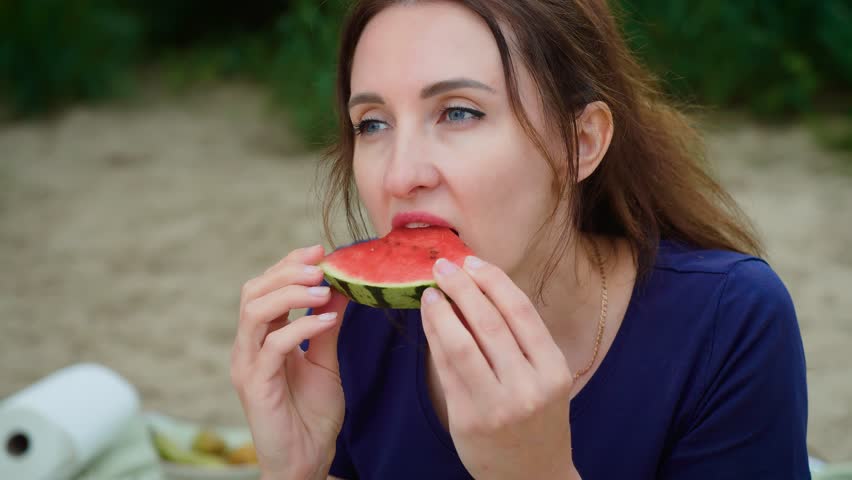 Woman eating watermelon with focused expression and calm demeanor. Female biting watermelon slice while showing neutral face. Lady consuming fruit with thoughtful gaze