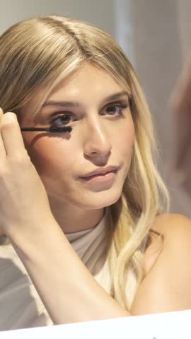 Reflection of crop young transgender woman using mascara applicator on eyelashes while doing makeup in mirror at bathroom