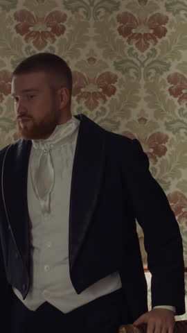 Vertical medium shot of noble Caucasian young man in formal attire standing by window in elegant room with classic decor, reading handwritten love letter then holding it close to his chest and smiling