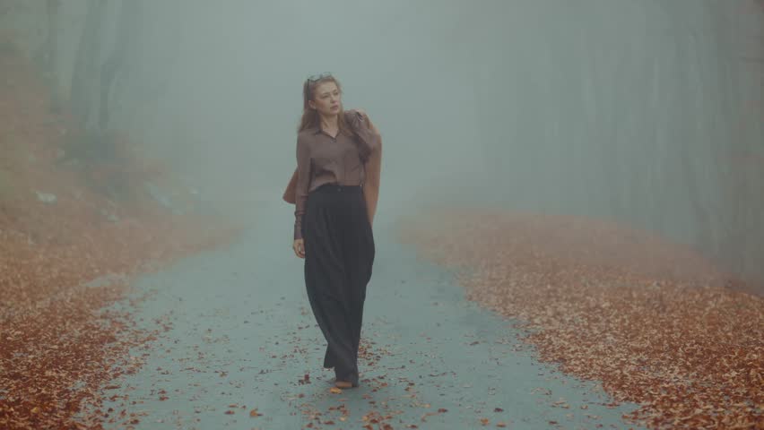 A woman walks thoughtfully along a misty path covered in fallen leaves, surrounded by serene nature in autumn.
