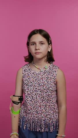 Vertical portrait of serious gen alpha girl with prosthetic arm stretching rubber band of beaded necklace and looking at camera standing against pink wall