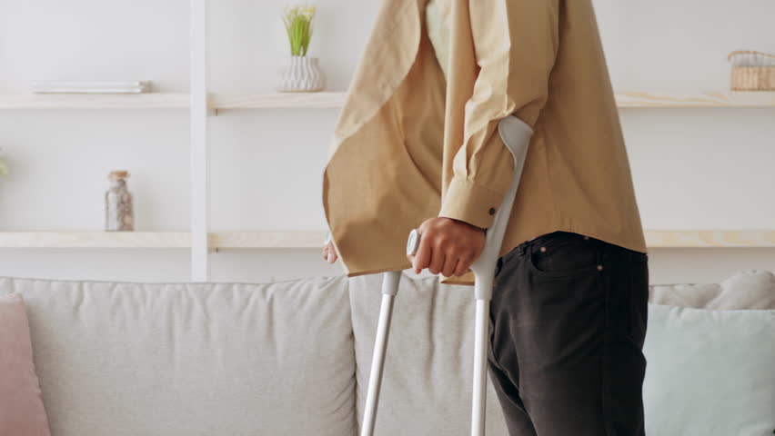 A millennial man with a disability stands in his living room, using crutches for support. The cozy home environment features a couch and minimal decor, creating a warm atmosphere.