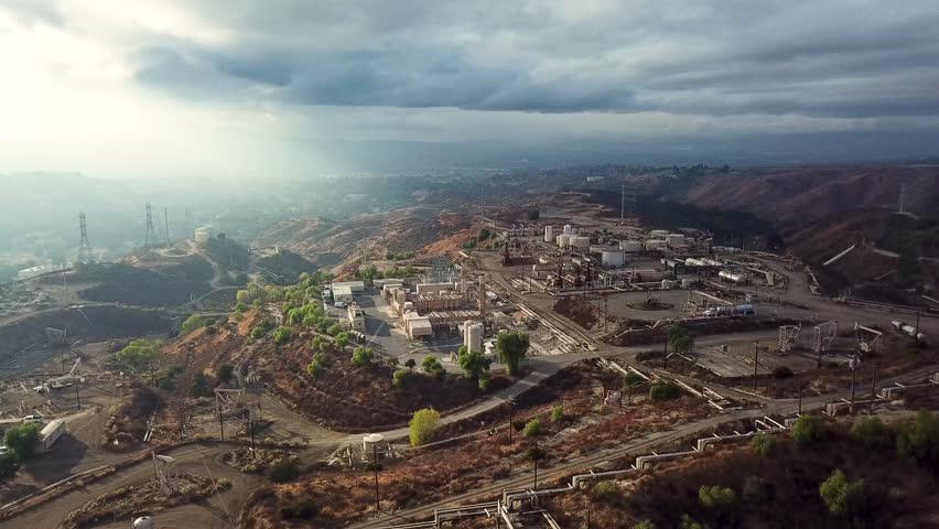 Aerial wide shot of oil factory on top of Californian mountains. Cloudy day with sunlight in USA. Pipelines and oil wells.