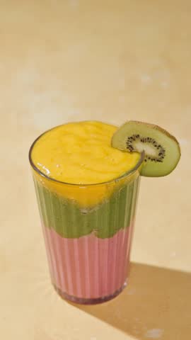 hand puts a straw into a glass of freshly made colorful spinach and banana mango and strawberry smoothie on a sunny light table surface