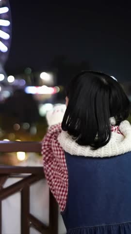 A Taiwanese woman in her 20s looks at the illuminated Ferris wheel from the stairs of an amusement park at night in Shilin District, Taipei City, Taiwan. Vertical slow motion video.