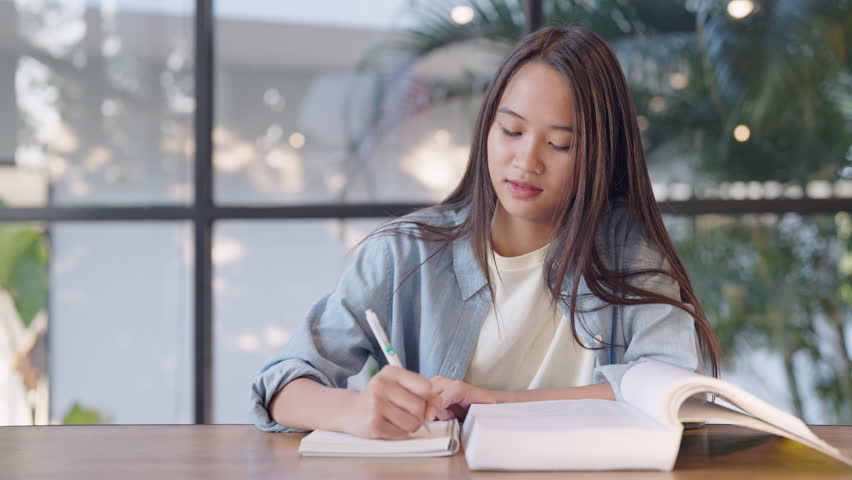 A woman is writing in a book. She is wearing a blue shirt and a white t-shirt