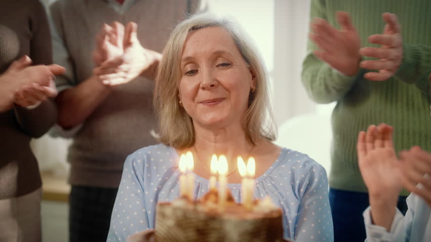 Charming elderly woman blowing candles on traditional birthday cake, slow motion . Happy old woman celebrating her anniversary with family, change of age and aging process, retiree female person