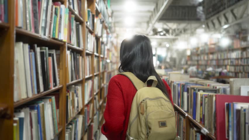 Young woman with a backpack browsing shelves in a cozy bookstore. Calm university student in a red sweater searching for a book in a quiet, well-lit library.