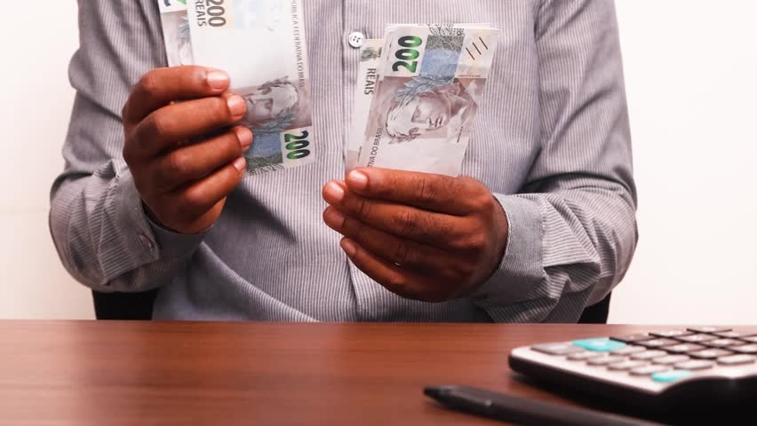 Black African male business man in long sleeves shirt sitting and counting Brazilian real notes with calculator and pen on desk
