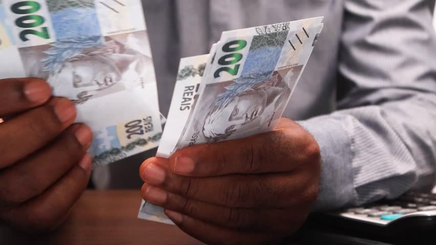 Black African male business man in long sleeves shirt sitting and counting Brazilian real notes with calculator on desk. close up

