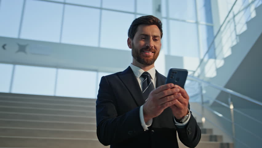 Smiling employee looking smartphone on staircase modern office building closeup. Confident executive in suit enjoy online communication by cellphone. Corporate manager browsing mobile phone content.
