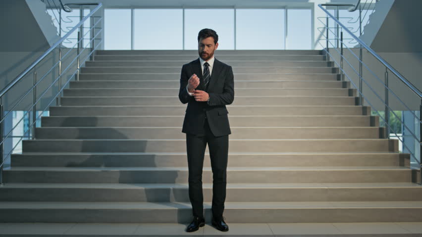 Focused businessman adjusting suit on indoors staircase. Confident executive preparing for business meeting in modern office. Successful ceo smiling to camera expressing determination standing stairs.