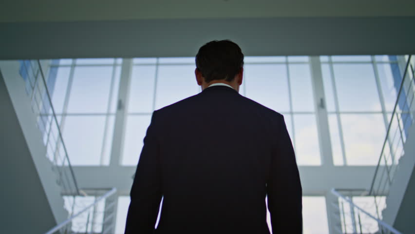 Elegant entrepreneur climbing stairs in bright modern office back view. Professional male executive moving upward with focused ambition in light interior. Corporate leader ascending stairway in suit.