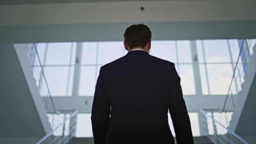 Determined businessman walking upstairs in light modern office back view. Stylish male professional climbing glass stairway confidently in elegant suit. Unknown executive man ascending staircase.