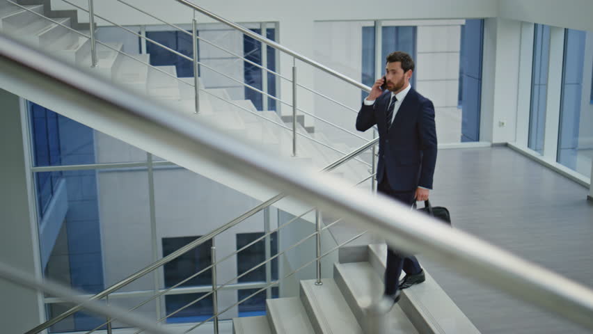Professional walking down stairway talking cellphone. Successful businessman calling smartphone going downstairs modern office building. Busy leader checking wristwatch descending stairs with bag.
