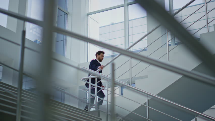 Ambitious manager climbing stairs inside bright modern office back view. Businessman in elegant suit walking upstairs hurrying on work. Confident entrepreneur going up staircase corporate building.