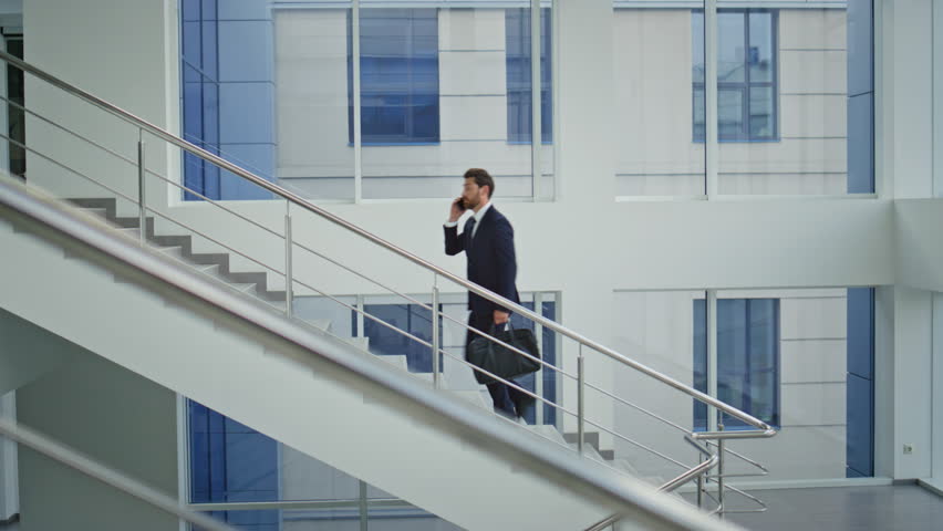 Bearded manager calling mobile phone walking upstairs modern light building. Busy businessman talking cellphone climbing staircase with briefcase. Rich executive have business conversation on go.