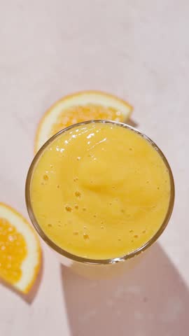 hand puts a straw into a glass of freshly made yellow orange and mango banana smoothie on a sunny light table surface