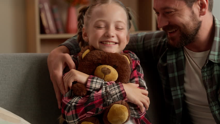 Pleased Caucasian child schoolgirl hug toy teddy bear happy dad daddy parent cuddling touch smiling daughter at home birthday gift man guy kid celebrate family father little girl enjoy holiday bonding
