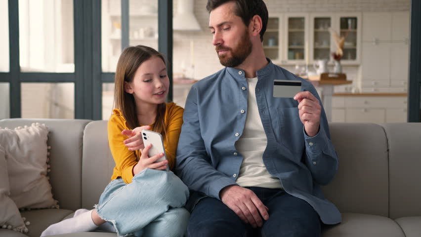 Joyful Caucasian parent man father holding credit card guy male talking finance smiling little schoolgirl girl child using mobile phone e-commerce at home family remote kid daughter shopping dad pay