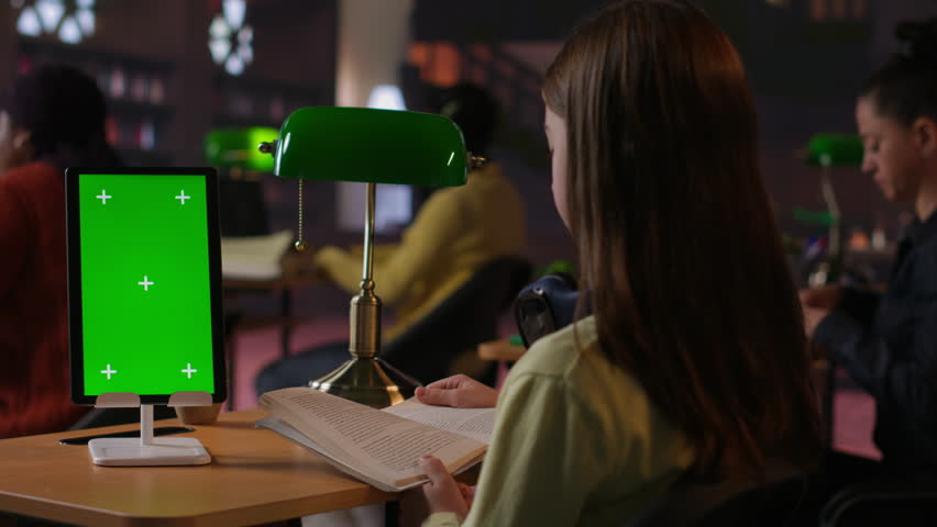 Schoolgirl reading a book next to copy space on tablet at the library desk, using academic database for research. Completing school assignments and developing her educational skills. Camera B.
