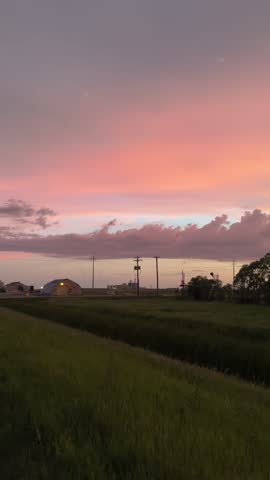 Industrial structures silhouette against a dramatic sunset with vibrant orange and purple clouds. Rolling grassy fields create a striking contrast.
