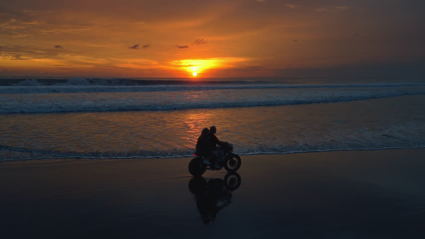 Couple of young woman and man on vintage motorcycle ride along the beach in evening. Male and female lead a scooter on the shore or coast of Bali, Indonesia. Road trip in exotic country. Drone aerial