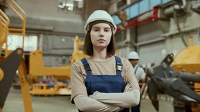 Female technician in hard hat and overalls holding arms crossed, posing confidently to camera in industrial factory, colleagues discussing machinery in background. Video portrait - Powered by Shutterstock - Get 15% off with code: PIKWIZARD15