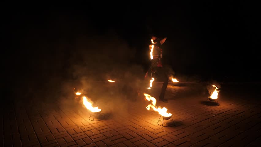The stuntman actress performs with fire torches at night outdoors. Bowls of kerosene are placed around the woman with burning torches, the fire is on show. A fire show with special effects.