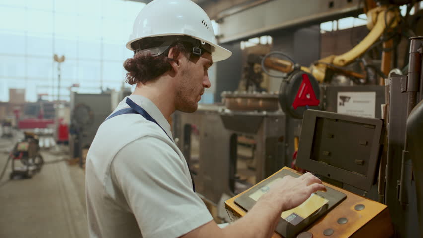 Male worker wearing safety hard hat and overalls, operating welding machine with control panel in industrial factory