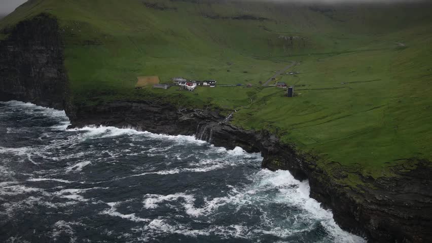 Stormy sea waves crashing against the rocky cliffs of Kalsoy small village nestled on the green hills, creating a dramatic and breathtaking scene in the Faroe islands, slow motion drone static shot
