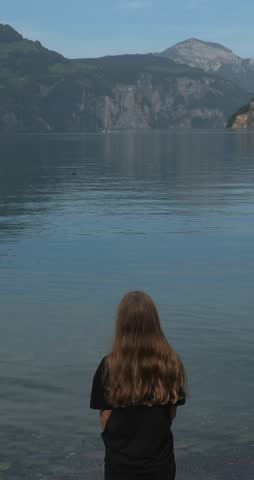 a young girl stay on stones on the shore of a lake in Switzerland and looks at the lake, vertical video content for social networks