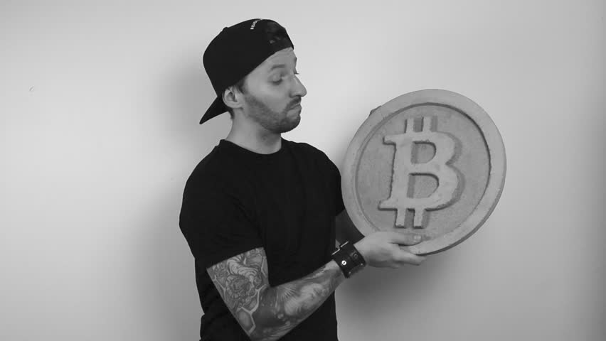 Grayscale footage of a tattooed man lifting up a large concrete or stone bitcoin sculpture and shaking his head in disapproval while not being happy. Cryptocurrency lifting up in slow motion by man.