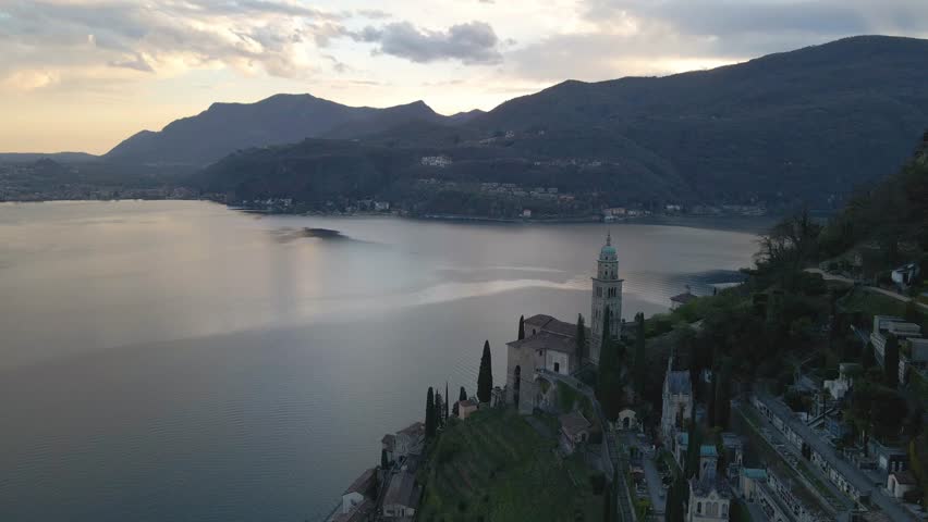 Morcote village of in Ticino, Switzerland, with its iconic church and cemetery, emerging from the morning mist on lake lugano as the sun rises over the majestic alps mountains drone pull out