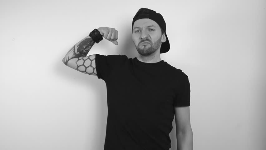 Grayscale footage of a tattooed man showing his bicep off by raising his arm and flexing his muscle in front of a white background. Man creates shadows on the wall behind him while doing expressions