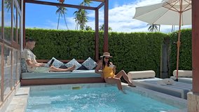 In a serene pool villa in Thailand, a dedicated freelancer works on his laptop while a companion enjoys leisure time by the water. A diverse couple of man and woman at a pool villa - Powered by Shutterstock - Get 15% off with code: PIKWIZARD15