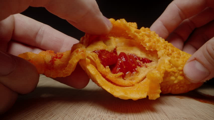 Fingers Gently Opening a Bitter Melon, Revealing Shiny Red Seeds Inside the Bumpy Orange Skin on a Wooden Surface.