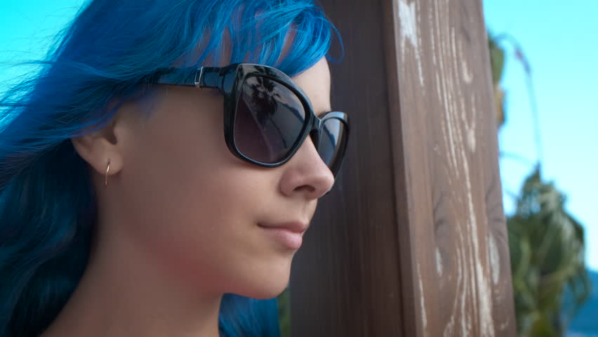 Stylish teen looks at the sea water. A stylish girl with blue hair and glasses relax on the beach under the sun light in summer.