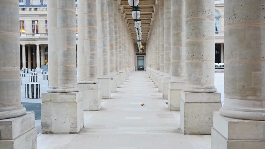 A Parisian woman in a grey beret and coat walks near the classic stone columns of Palais Royal, showcasing timeless French elegance.