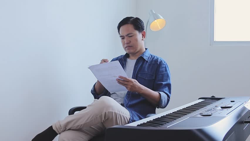 Asian Male Songwriter Writing A Song On Music Sheet While Sitting By Electronic Keyboard In Home Studio