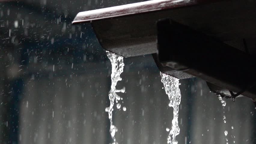 A rain gutter is dripping water. The water is falling from the gutter and splashing onto the ground. The scene is dark and moody, with the rain creating a sense of melancholy