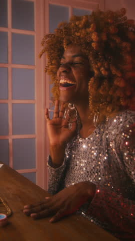 Vertical side lowkey shot of 70s young Black female singer with puffy hair in glittery silver dress, warming up her voice and enjoying herself, showing off in front of vanity mirror in dressing room