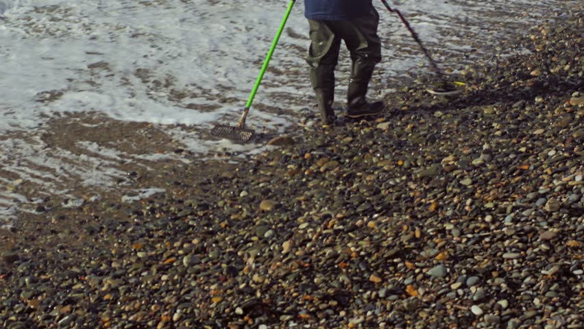 Close-up of a man in high rubber boots and jacket. He is holding a metal detector and a shovel. The man is looking for precious jewellery on a rocky seashore. Searching for gold on seashore