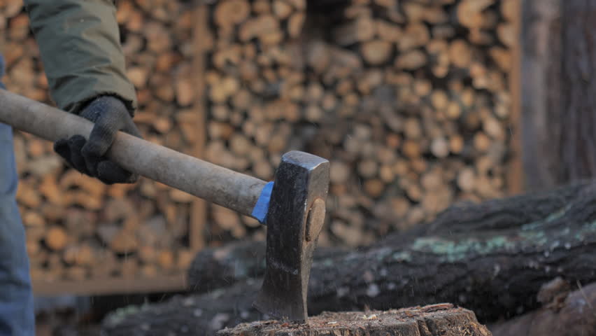 An axe strikes a solid log resting on a sturdy stump, sending splinters flying. In the background, stacked firewood creates a cozy outdoor atmosphere.
